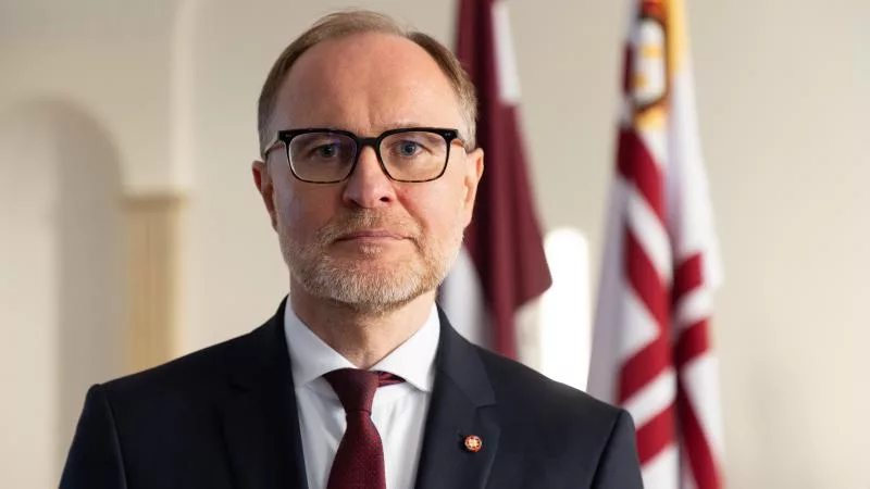 Öltönyös, szemüveges férfi zászlók előtt