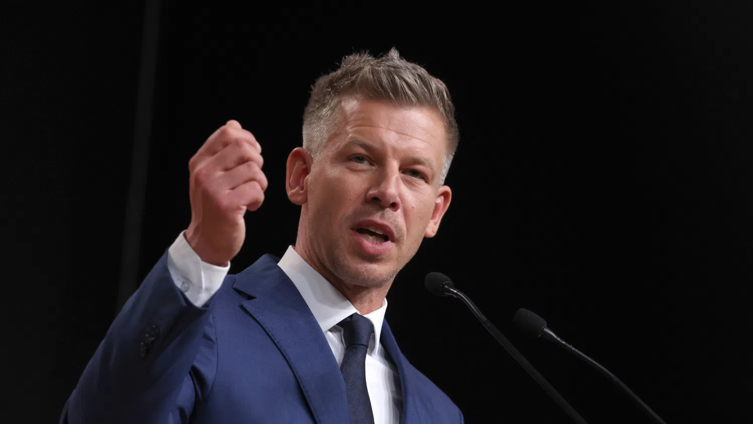 Kék öltönyös férfi beszédet mond mikrofon előtt